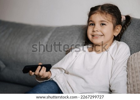 Portrait of cute little girl using remote controller of TV and watching cartoons. Small but already so grown up