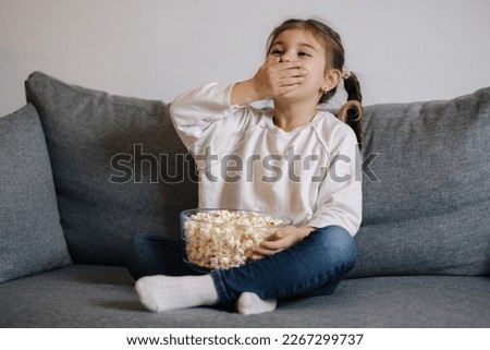 Little girl watching a cartoon emotionally and covers her mouth with hand in surprise. Popcorn. Holiday mood