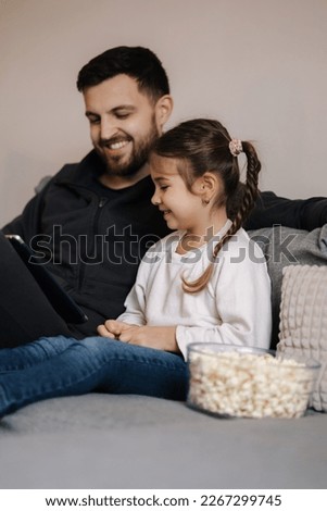 Adorable little girl watching cartoons on table with her father. Daughter with dad eating popcorn and laugh. Home mood