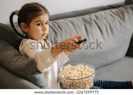 Cute little girl using remote controller of TV and watching cartoons. Four year girl eating popcorn on sofa