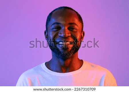 Closeup portrait of cheerful handsome bearded middle aged black man in white posing on colorful neon studio background, smiling at camera. People faces, human emotions concepts