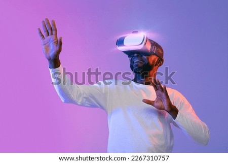 African american man in white experiencing virtual reality over neon studio background, using modern wireless VR goggles, moving hands, playing video game, copy space