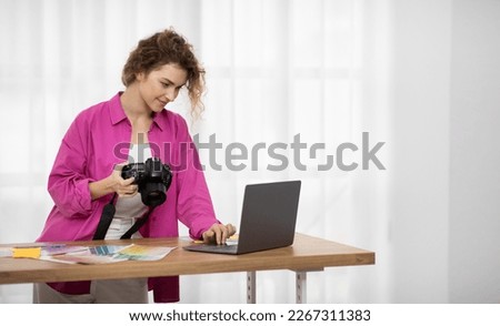 Beautiful Female Photographer Holding Photocamera And Using Laptop Computer While Working At Home Studio, Smiling Woman Standing At Desk Near Window And Processing Images On Computer, Copy Space