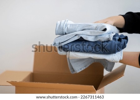 Closeup Shot Of Woman Packing Stack Of Clothes In Cardboard Box, Unrecognizable Female Putting Pile Of Jeans To Parcel, Ready To Make Donation Or Sell Used Clothing, Decluttering Home, Cropped Image Royalty-Free Stock Photo #2267311463