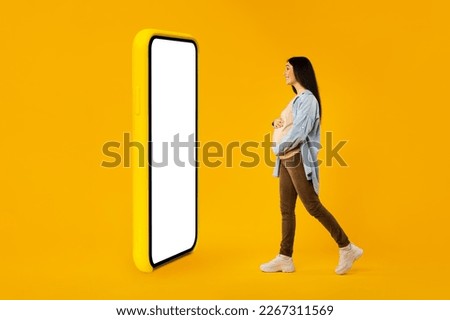 Happy pregnant woman walking to big smartphone with blank screen, expectant lady advertising new app or website over yellow background, creative collage, mockup