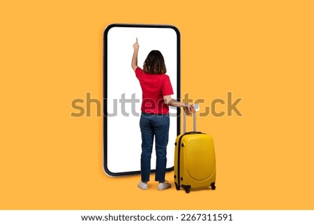 Back view of brunette woman tourist with yellow luggage using big smartphone with white blank screen over yellow studio background, booking hotel or airplane tickets online, mockup, copy space Royalty-Free Stock Photo #2267311591