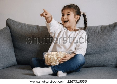 Adorable girl watching a cartoon emotionally and points his hand in the direction of the TV. little girl eating popcorn