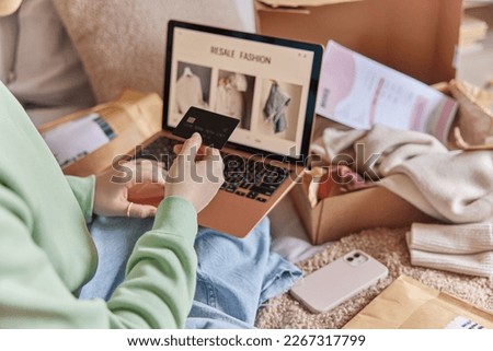 Cropped image of unrecognizable woman holds credit card and uses laptop computer makes shopping online buys clothes in internet webstore from home. Focus on banking card. Close up. Resale fashion