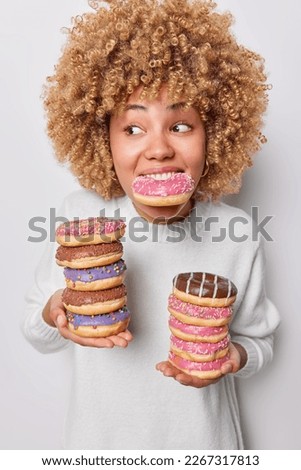 Vertical shot of curly haired woman poses with stack of appetizing glazed doughnuts breaks diet eats harmful high calories food dressed in casual jumper focused aside isolated over white background Royalty-Free Stock Photo #2267317813
