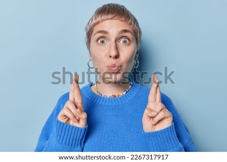 Hopeful short haired woman keeps lips rounded crosses fingers believes in good luck wears casual jumper earrings and necklace isolated over blue background wishes for good luck prays for something Royalty-Free Stock Photo #2267317917