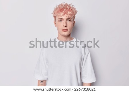 Portrait of attentive hipster guy with pink hair and tattoo on arm dressed in casual basic t shirt looks directly at camera has serious expression isolated over white background belong to subculture Royalty-Free Stock Photo #2267318021