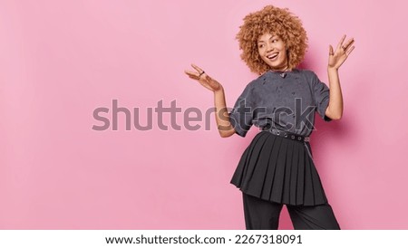 Horizontal shot of cheerful curly haired woman keeps palms raised up smiles broadly dressed in t shirt skirt foolishes around has happy mood isolated on pink background blank space for your promotion