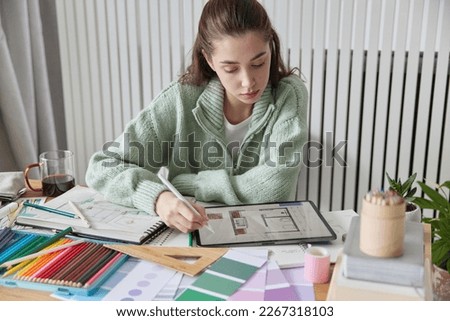 Indoor shot of young female interior designer works on apartment design project draws in digital tablet with stylus thinks about decorations of apartment wears casual jumper poses at desktop. Royalty-Free Stock Photo #2267318103