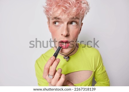 Photo of pink haired gay applies lipstick wears makeup wears feminine clothes prepares for date has women appearance isolated on white background. Non conforming queer man poses with cosmetic product
