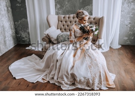 A beautiful blonde bride in a white lace dress sits on a beige sofa with a bouquet of flowers in a retro studio on a gray background. Wedding photography, portrait. Royalty-Free Stock Photo #2267318393
