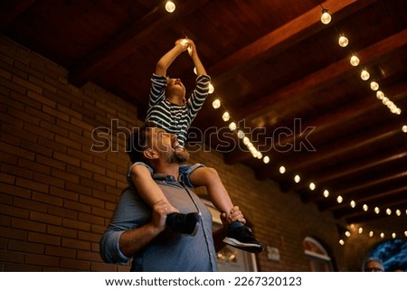 Happy father carrying his son on his shoulders while turning on the lightbulb on a patio. Royalty-Free Stock Photo #2267320123