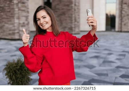 Close-up portrait of a young happy woman, holding a smartphone in one hand, thumbs up with the other hand.