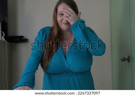 a pregnant woman in a blue dress puts her hand to her face in a face palm gesture as she is embarrassed by a mistake she made. She seems to be having fun and making light of the moment. Royalty-Free Stock Photo #2267337097