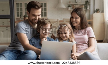Happy young caucasian family with small children relax on sofa in kitchen have fun using laptop together, smiling parents rest on couch enjoy domestic weekend with little kids watch video on computer