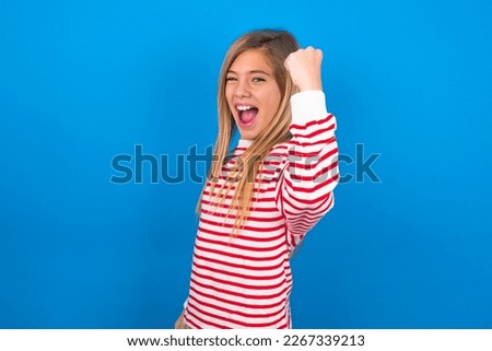 Overjoyed Beautiful caucasian teen girl wearing striped shirt over blue background glad to receive good news, clenching fist and making winning gesture.