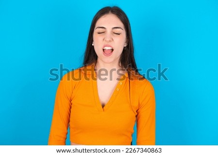 young woman wearing orange crop top over blue studio background yawns with opened mouth stands. Daily morning routine