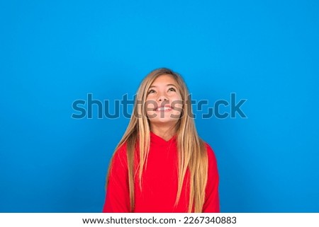 Portrait of mysterious Beautiful caucasian teen girl wearing red sweatshirt over blue background looking up with enigmatic smile. Advertisement concept.