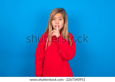 Beautiful caucasian teen girl wearing red sweatshirt over blue background makes hush gesture, asks be quiet. Don't tell my secret or not speak too loud, please!