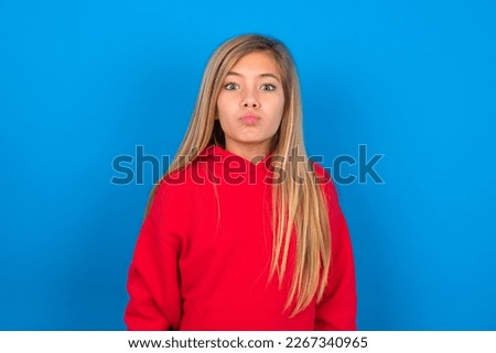 Beautiful caucasian teen girl wearing red sweatshirt over blue background, keeps lips as going to kiss someone, has glad expression, grimace face. Standing indoors. Beauty concept.