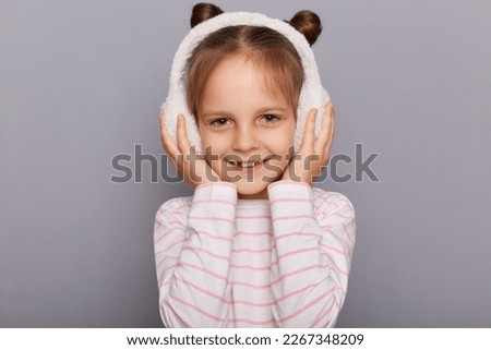 Indoor shot of smiling charming cute little girl wearing fur earmuffs and striped shirt standing isolated over gray background, looking at camera with toothy smile.