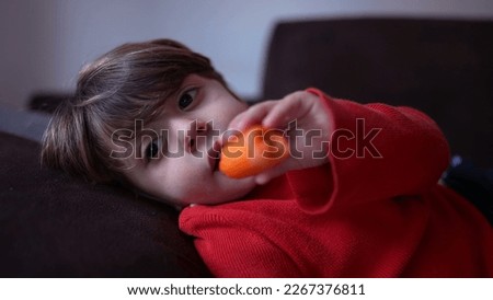 Child watching cartoon off camera lying on sofa couch at home while eating healthy snack carrot