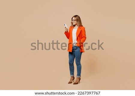 Full body young happy employee business woman corporate lawyer 30s wear classic formal orange suit glasses work in office hold in hand use mobile cell phone isolated on plain beige background studio