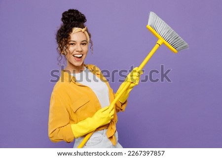 Side view young happy housekeeper woman wear yellow shirt rubber gloves tidy up hold broom sweeps floor clean house isolated on plain pastel purple background studio. Housework housekeeping concept