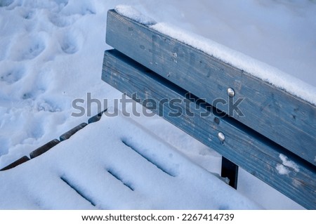 Light blue wooden bench covered by snow in a public park in Finland. Winter at park. Footprints in the snow. Simple bench. Cold weather. Outside photo.