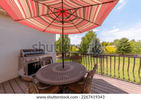 Terrace with dining table with umbrella and chairs near the gas griller. Dining table on a terrace with wooden flooring and metal railings against the lawn yard with trees against the sky.
