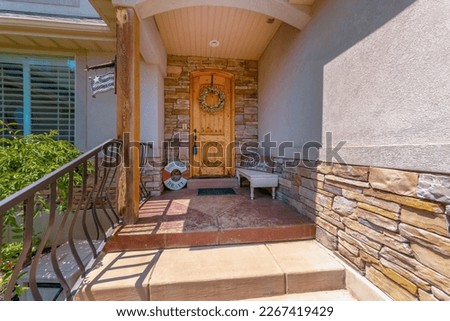 Arched wood front door with wreath at the entrance of the house with stone veneer and beige wall. Decorative house porch with railings and window on the left. Royalty-Free Stock Photo #2267419429