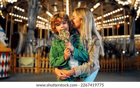 A cute caucasian boy with blonde curly hair with his mother eating a colorful lollipop standing against the background of a carousel with horses in the evening at an amusement park or circus.