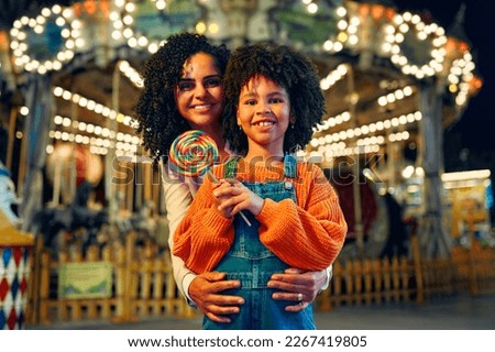 A cute African-American girl with an Afro hairstyle with her mother eating a colorful lollipop standing against the background of a carousel with horses in the evening at an amusement park or circus. Royalty-Free Stock Photo #2267419805