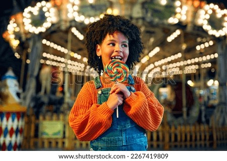 A cute African-American girl with an Afro hairstyle eats a colorful lollipop standing against the background of a carousel with horses in the evening at an amusement park or circus. Royalty-Free Stock Photo #2267419809
