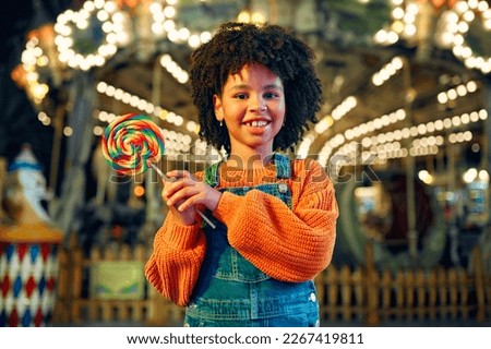 A cute African-American girl with an Afro hairstyle eats a colorful lollipop standing against the background of a carousel with horses in the evening at an amusement park or circus. Royalty-Free Stock Photo #2267419811