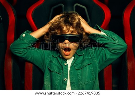 A cute Caucasian boy with curly hair sitting in a virtual cinema in glasses on moving rotating chairs and emotionally watching a movie in an amusement park.