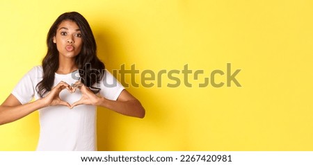 Portrait of beautiful african-american girl, pouting for kiss and showing heart sign, like or love someone, standing over yellow background.