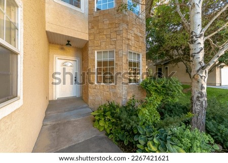 House entrance with plants and trees on the side heading to the white front door. There is a stucco wall on the left with window beside the entryway and bay windows with stone veneer on the right. Royalty-Free Stock Photo #2267421621