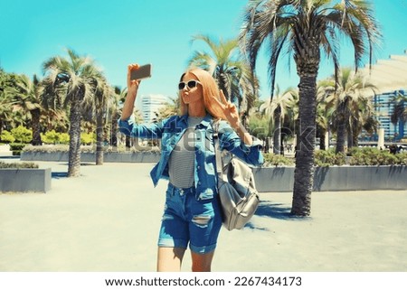 Portrait of young woman taking selfie with phone in summer park wearing backpack on palm tree background