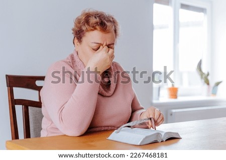 Tired and Sad Senior Woman at Home Struggling with Fatigue While Reading a Book. An old woman sits at home, reading a book with tired eyes and a look of fatigue on her face. Royalty-Free Stock Photo #2267468181