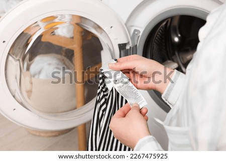 Woman reading clothing label with care symbols and material content on striped shirt near washing machine, closeup