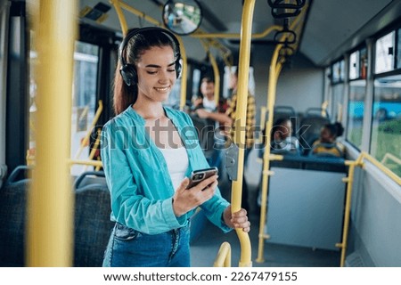 Young woman smiling while standing by herself on a bus and listening to music on a smartphone. Happy female passenger using headphones and a mobile phone in public transportation. Copy space. Royalty-Free Stock Photo #2267479155