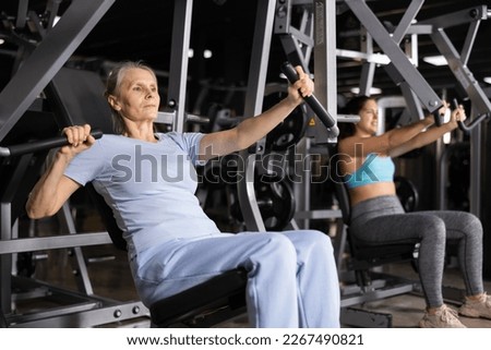 Concentrated aged woman leading healthy active lifestyle doing strength training in gym, performing chest press in exercise machine Royalty-Free Stock Photo #2267490821