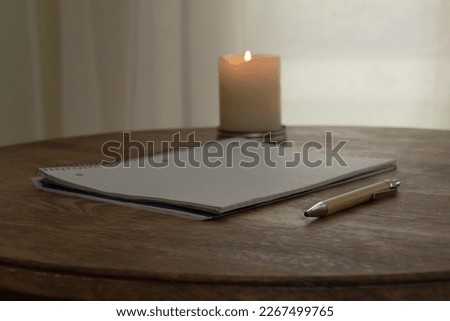 Desk with notebook, pen, and candle in soft, dark, and natural light.