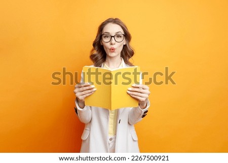 young surprised girl student in glasses and white suit holds book and is surprised on colored isolated background, female teacher reads yellow book in shock