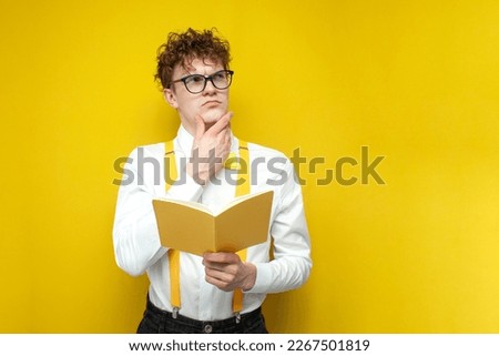pensive nerd guy in glasses holds book and thinks on yellow isolated background, student in festive outfit reads book and looks up at copy space
